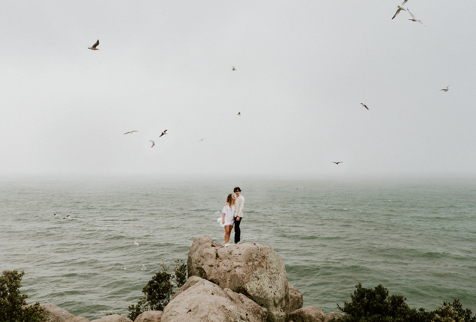 couple on top of rocks surrounded by flying birds with the ocean in the background