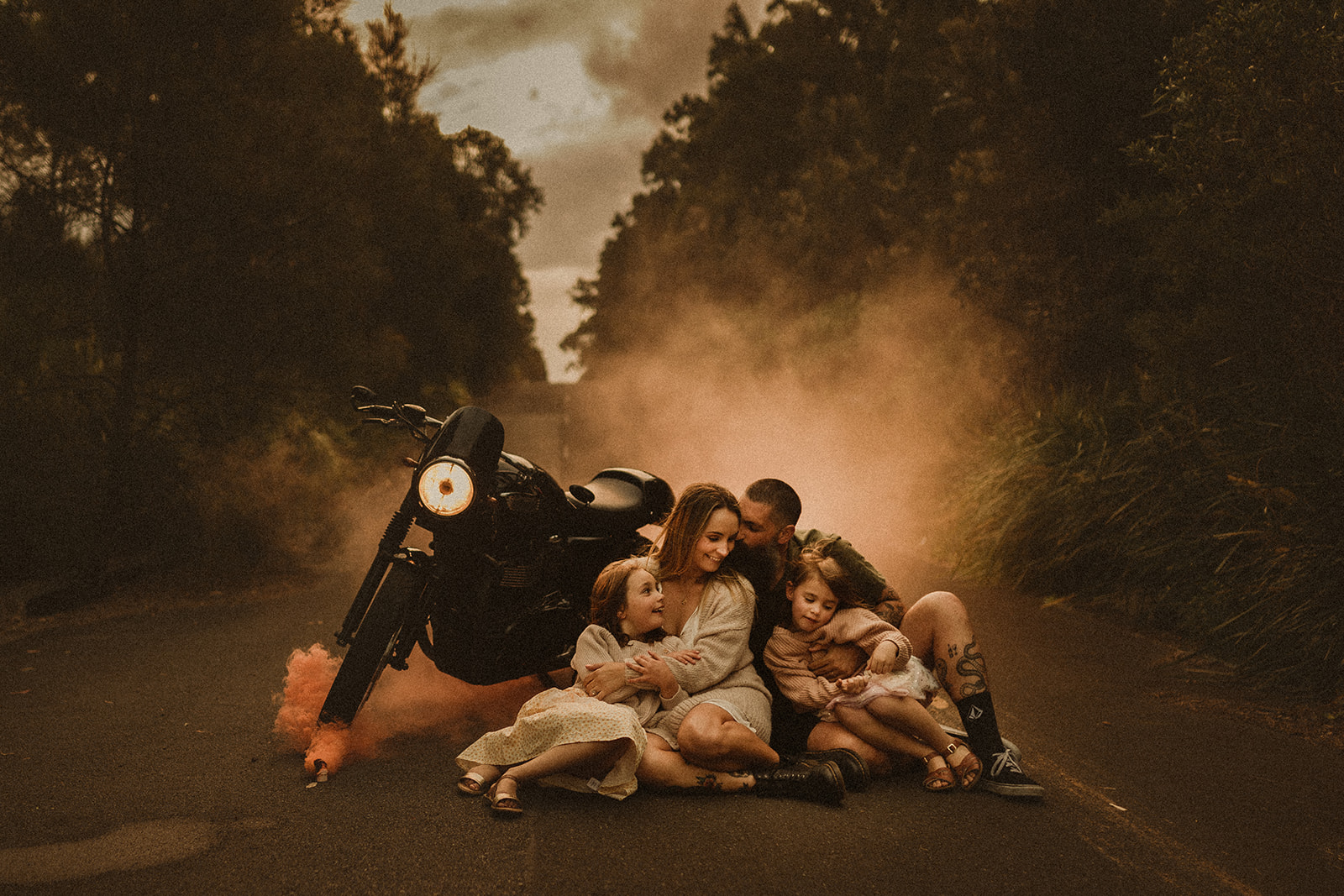 family cuddling near motorbike with orange smoke at dusk. Sydney Family Photography