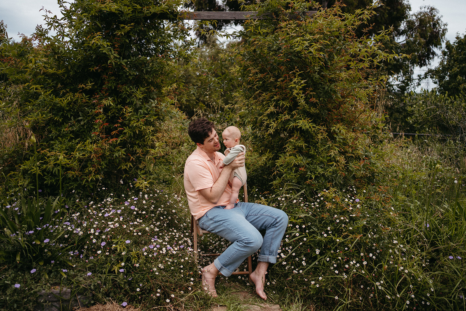 a father sits outsides in an overgrown flower thicket he is holding his young son on this lap