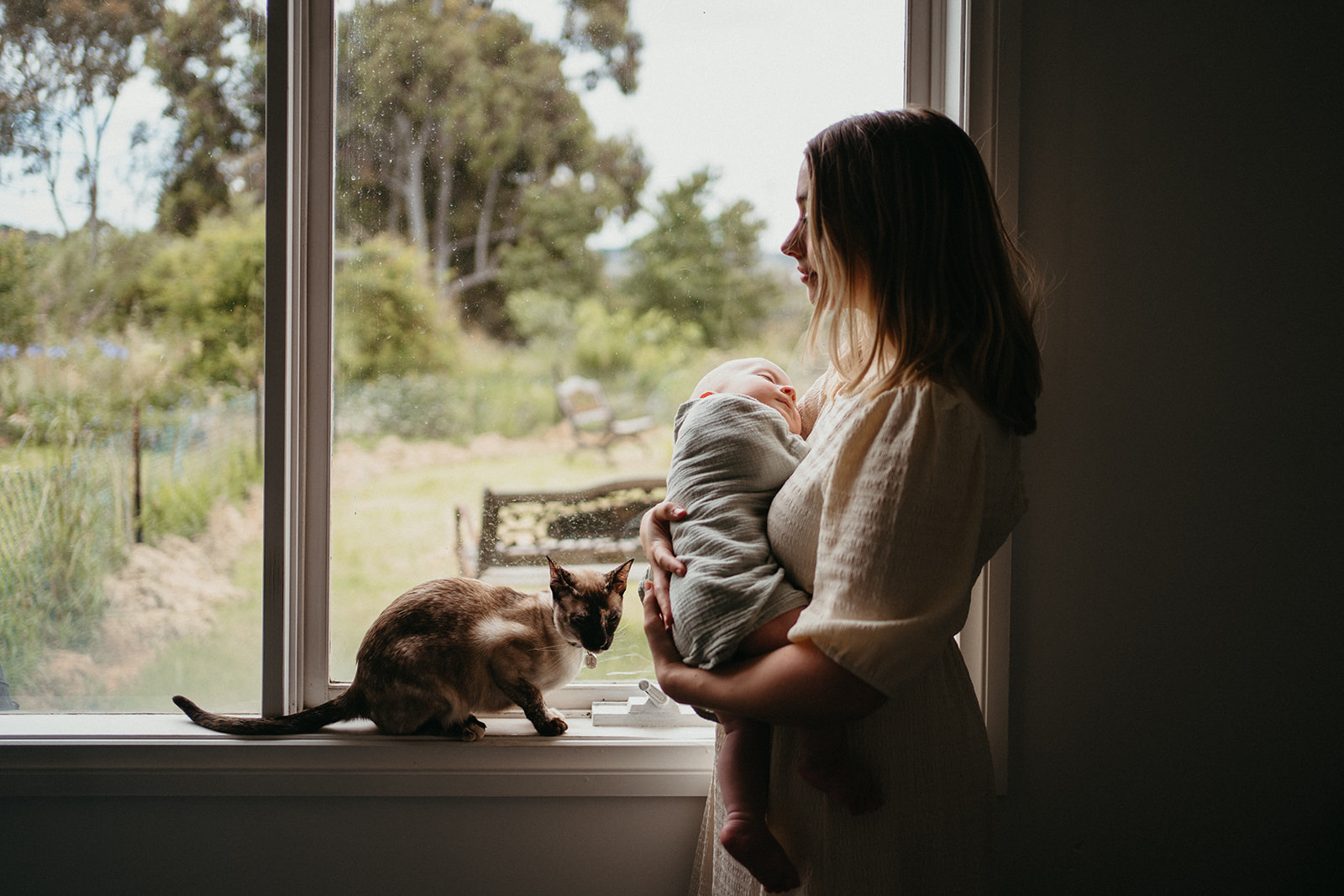 a mother holds her baby near a window a cat sits perched next to them