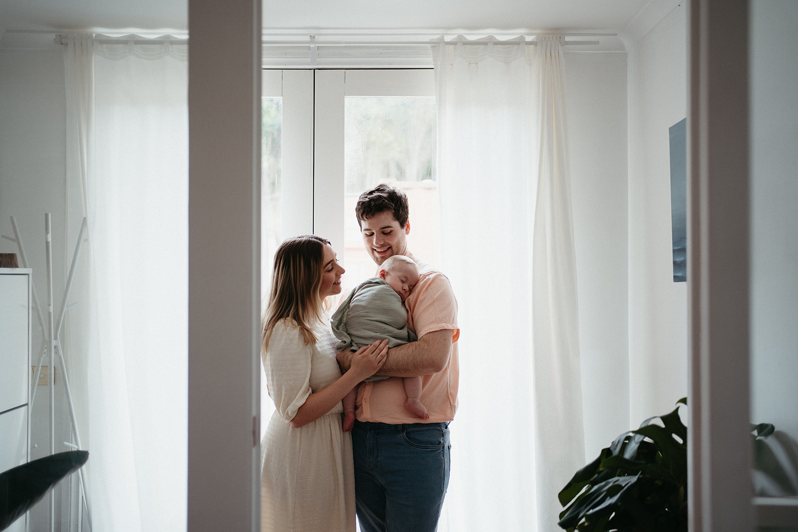 a young couple stand in a white room with curtains behind them they are holding their baby