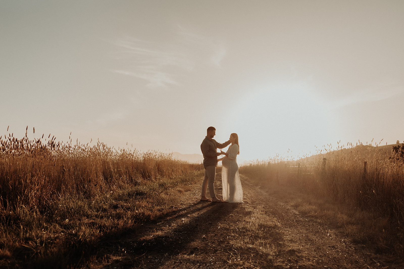 A couple who are about to have a baby stand on a dirt road, next to a grass field with the sunsetting behind them. 