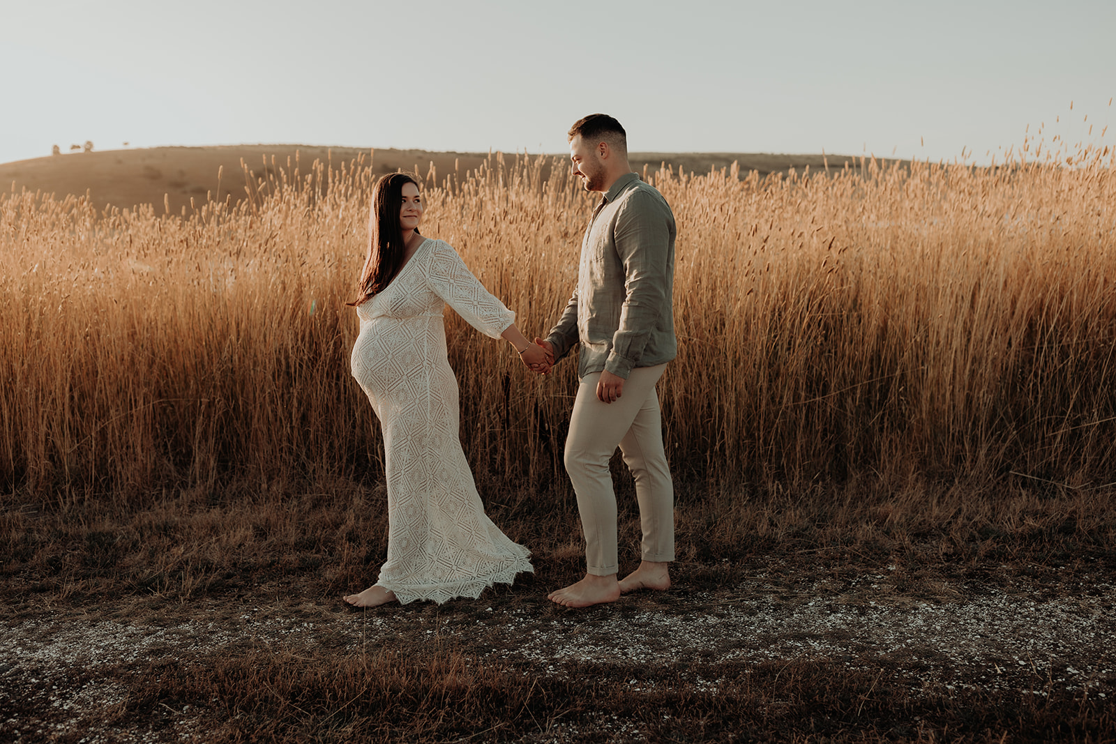 A heavily pregnant woman leads her partner by the hand past a field of tall grass. 