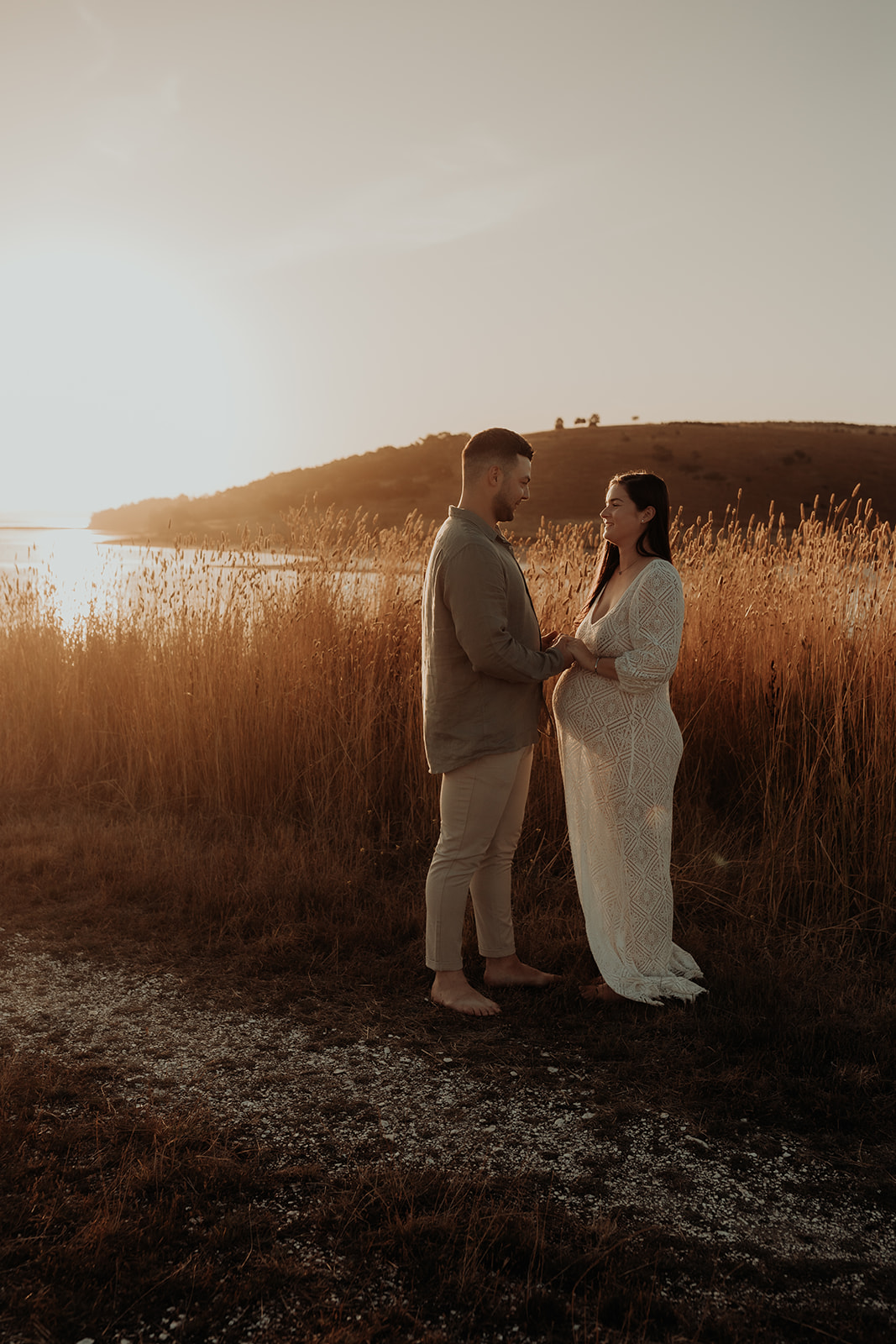 A man looks at his pregnant partner and touches her belly.  They are next to a grass field at sunset. 