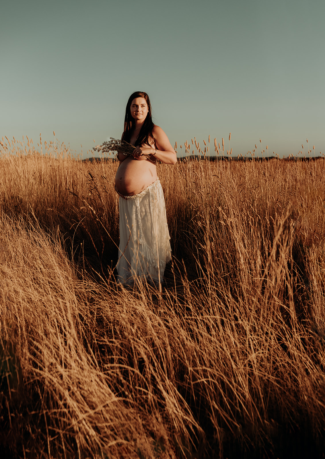 A heavy pregnant woman holds dried daisies to her naked chest, she wears a long white skirt and stands in a grass field.