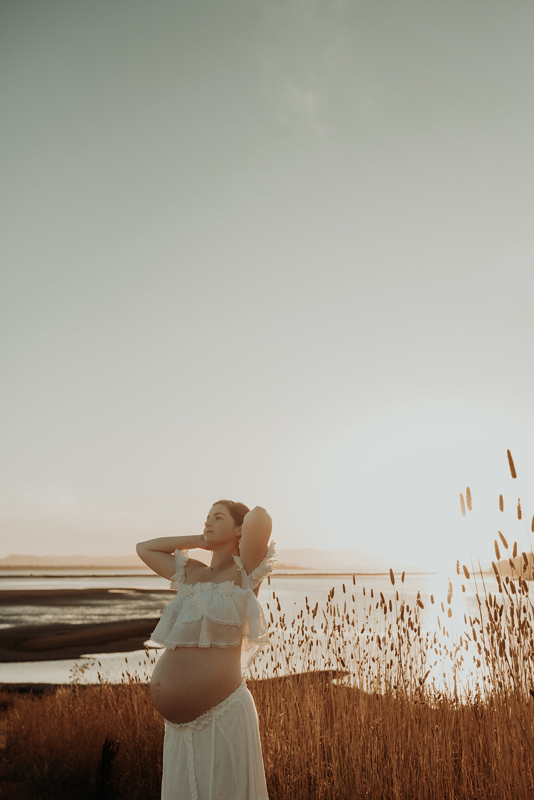 A pregnant woman stretches her arm above her head, next to a grass field, the sun sets over a bay behind her. 