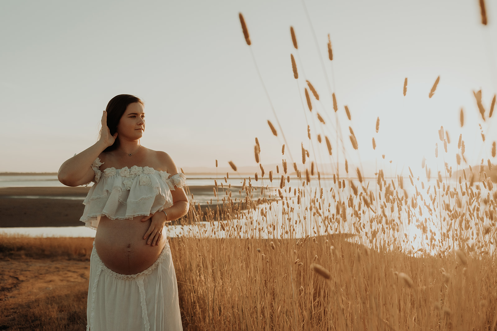 A pregnant woman next to a grass Field at sunset in front of a bay. 
