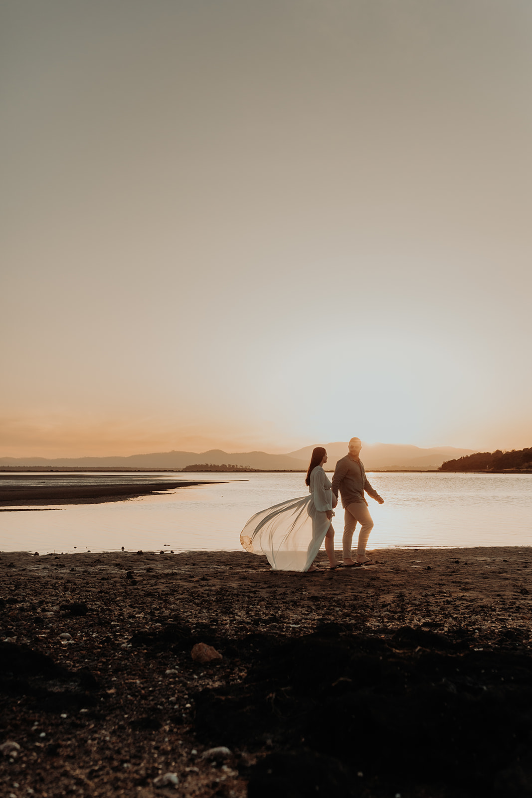 A man leads a pregnant woman along the shoreline of the baby at sunset. 