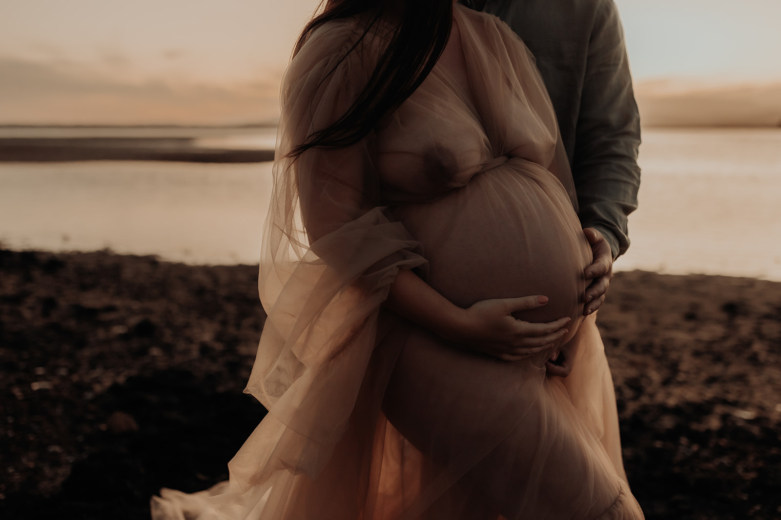 A man stands behind his pregnant partner and they both cup her belly with their hands.  Sunset over bay behind them. 