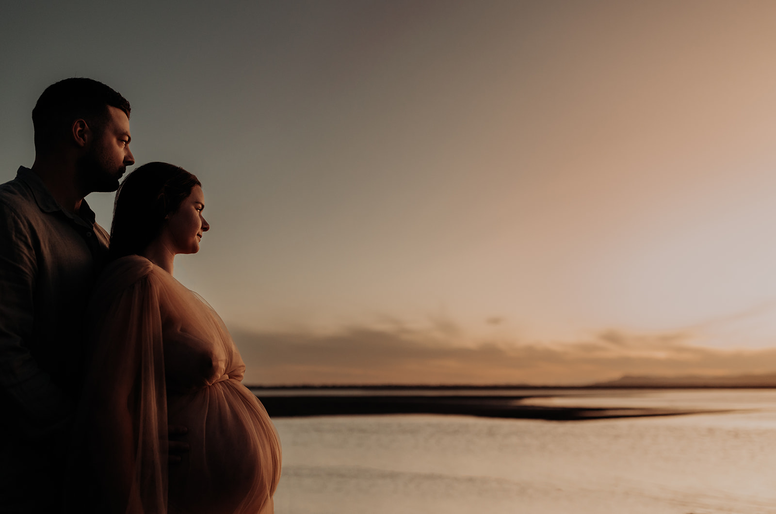 A pregnant couple look out towards the sun setting over the bay. 