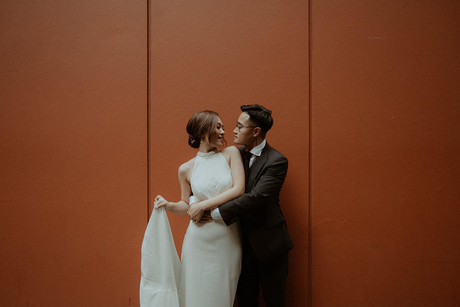 Bride and groom, hugging up against an orange wall
