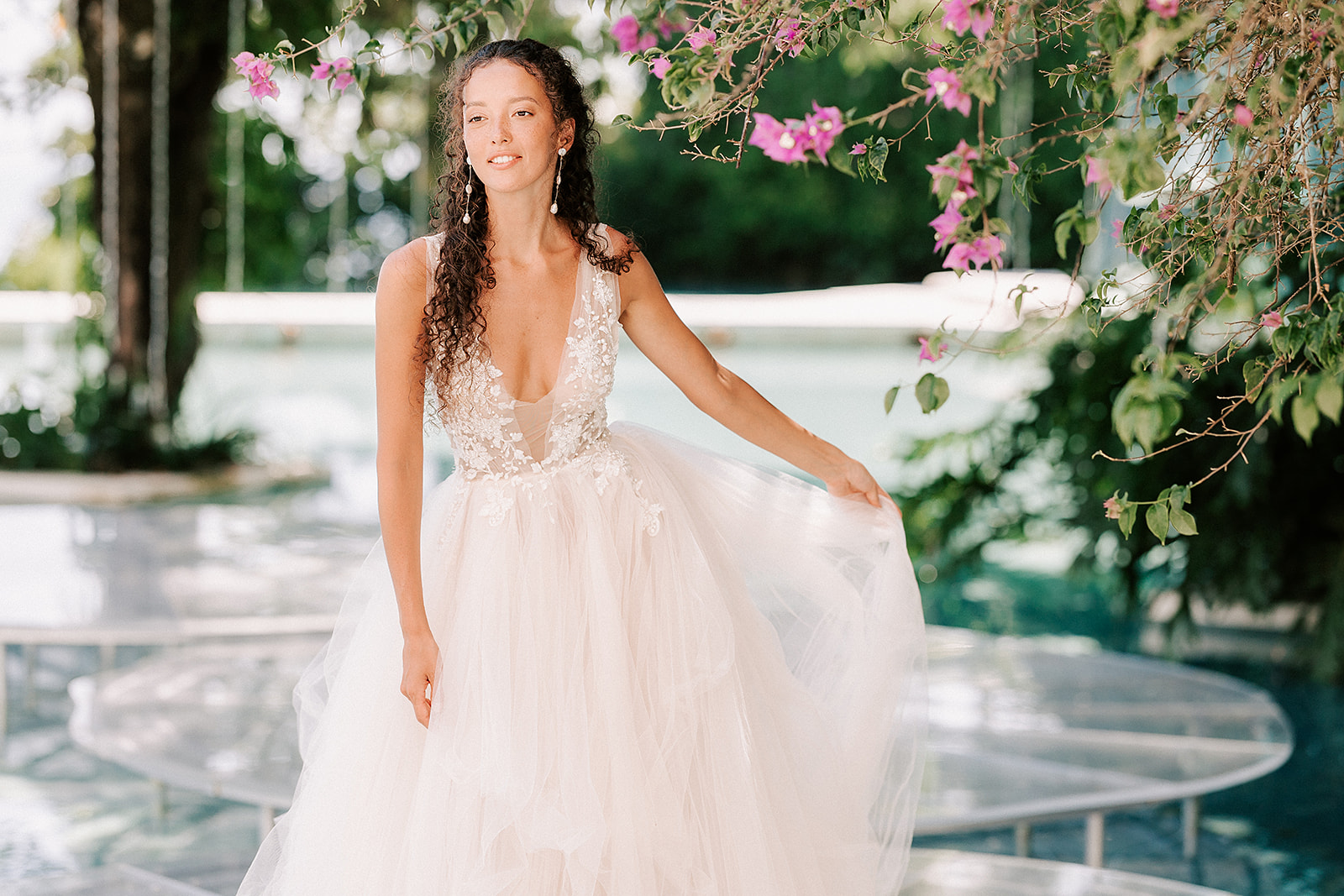 bride is holding the side of her dress with water in the background and pink flowers in the foreground