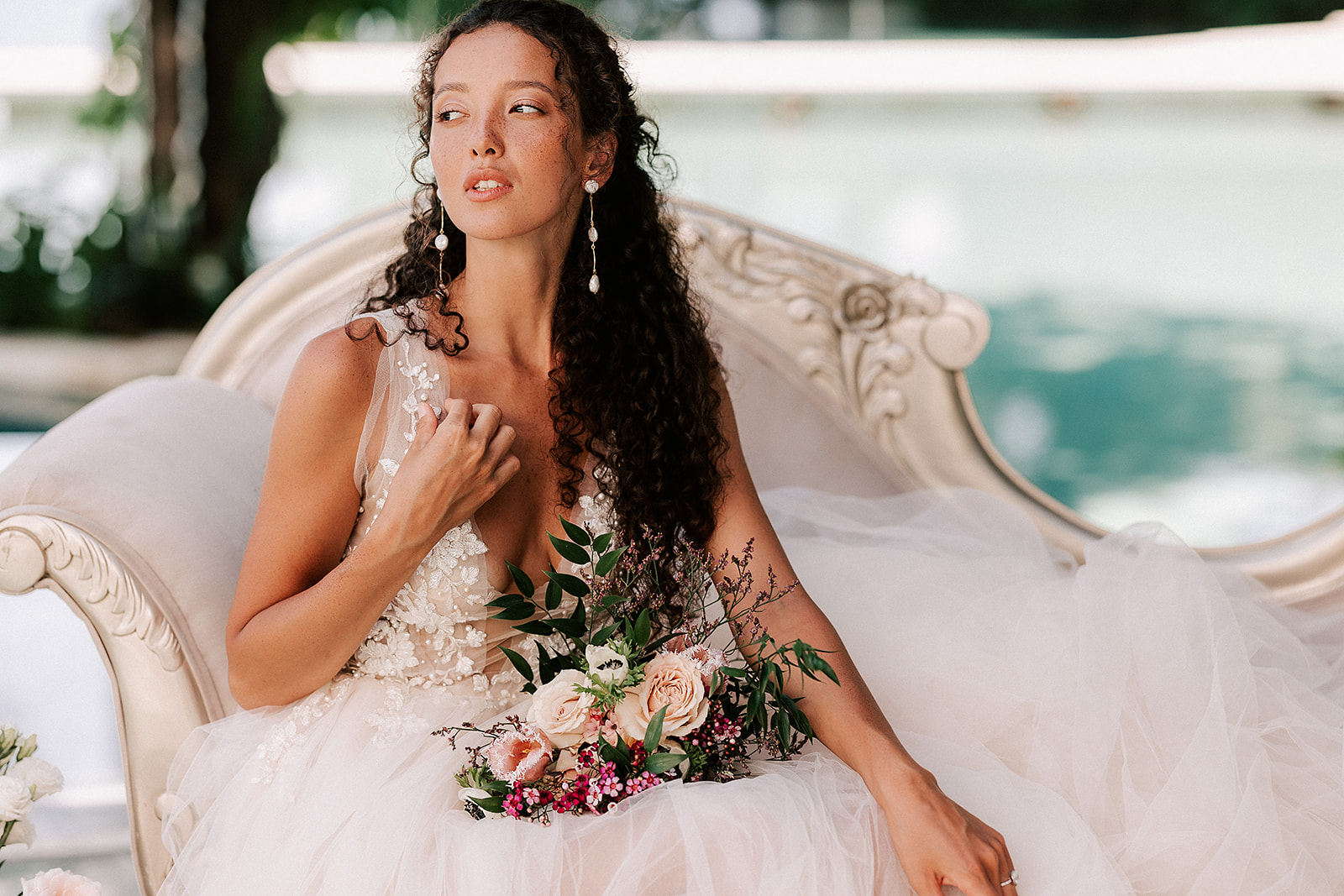Bride modelling on a couch with a bouquet on her lap and water in the background