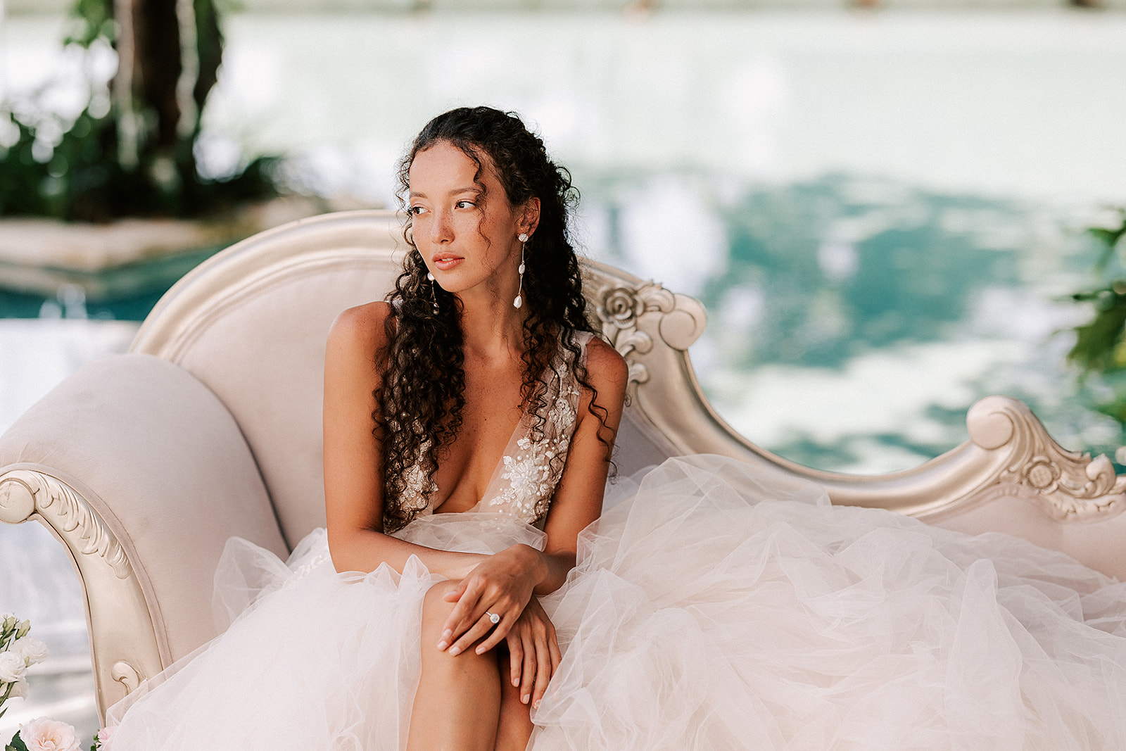 Bride is sitting on  a pink couch with legs crossed, and blue water in the background