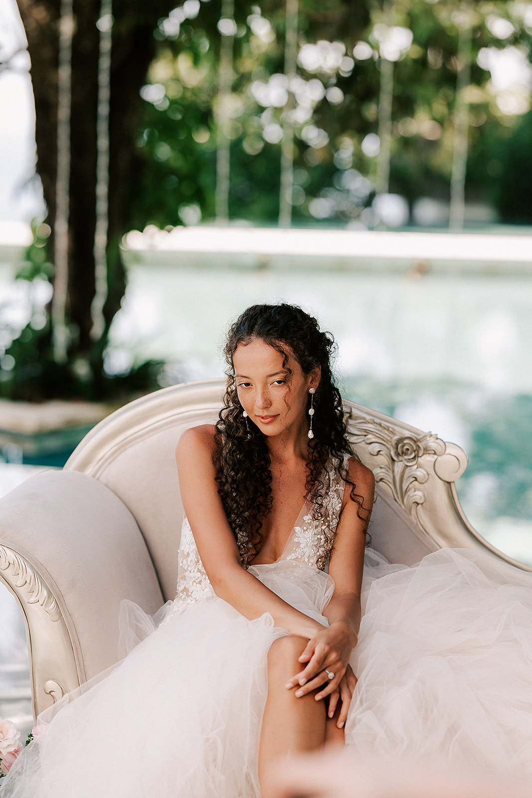 bride is sitting on a pink couch, outside with dreamy greens and florals in the background