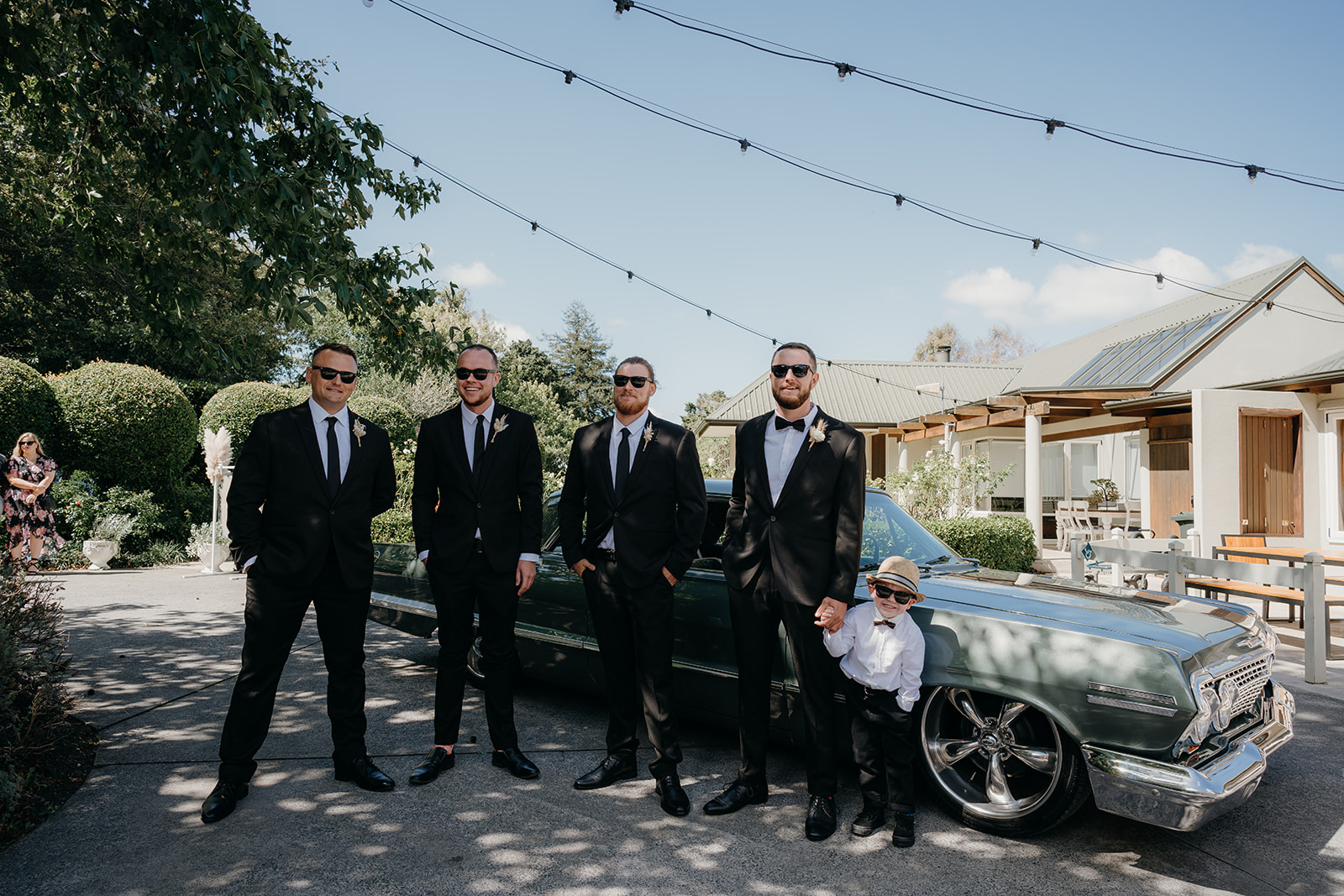 the groom and his groomsmen pose at tubala rasa in front of a vintage car under lights on a string

