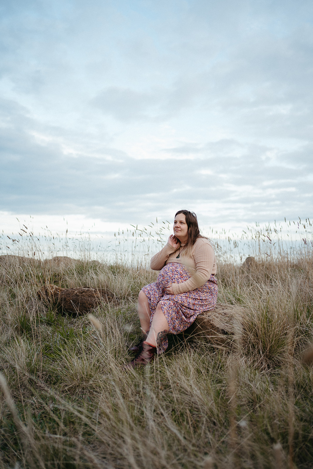 a pregnant woman sits on a rock in a field of grass, she is laughing