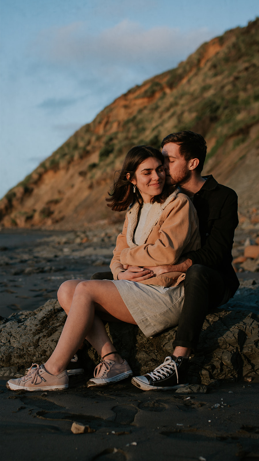 Port Waikato engagement portrait session. Couple sitting on a rock together hugging.