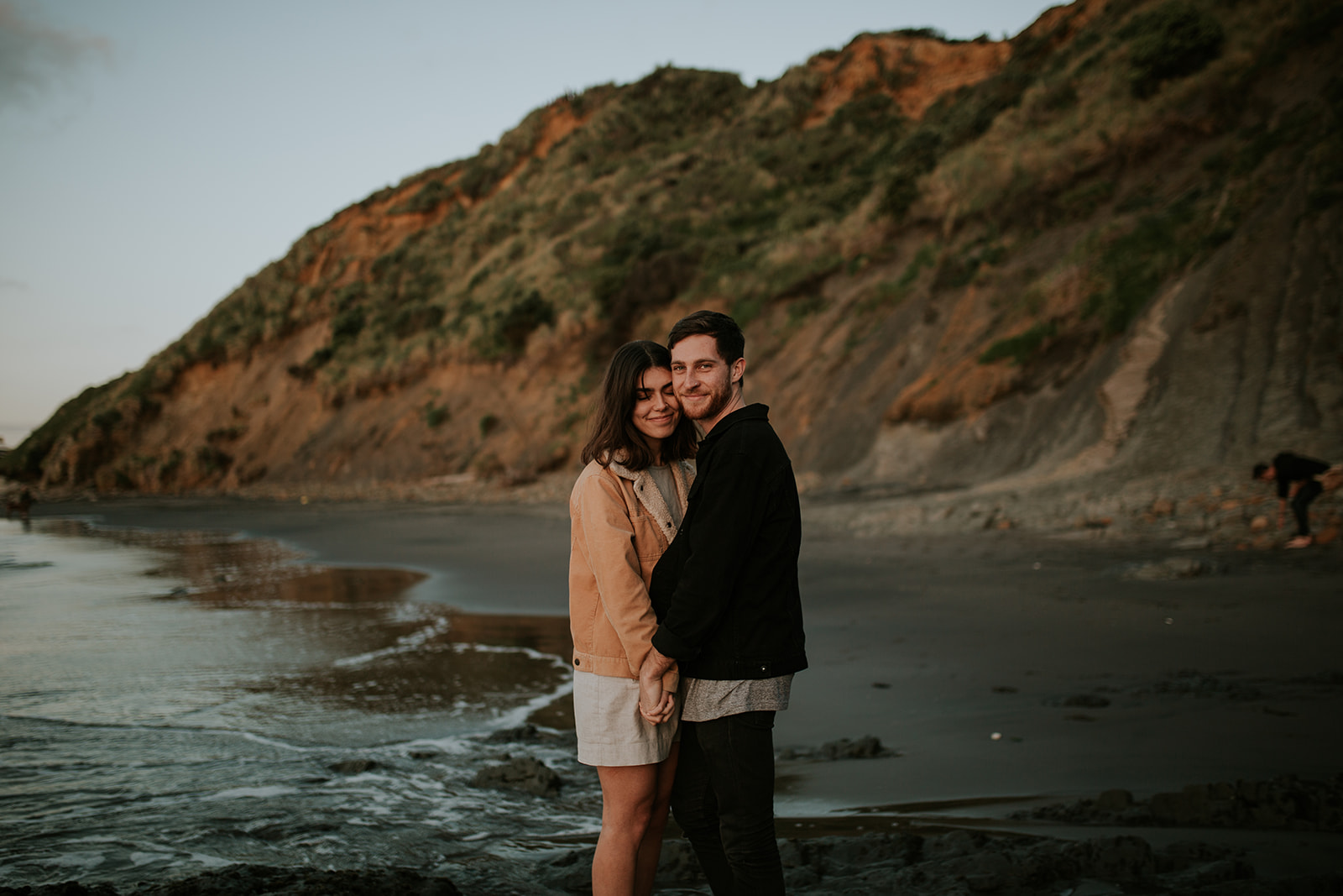 Port Waikato engagement session at sunset with couple standing in front of cliff faces.