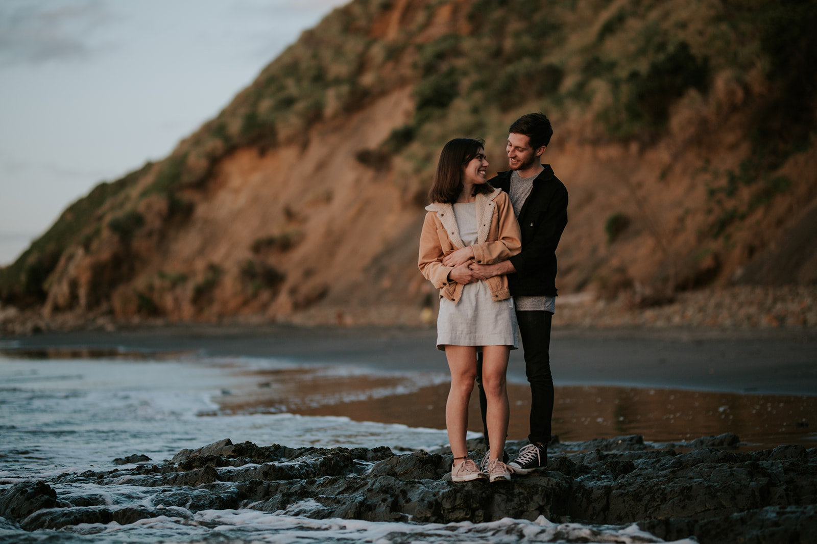 Sunset elopement at Port Waikato. Couple in casual attire standing on a rocks by the ocean.