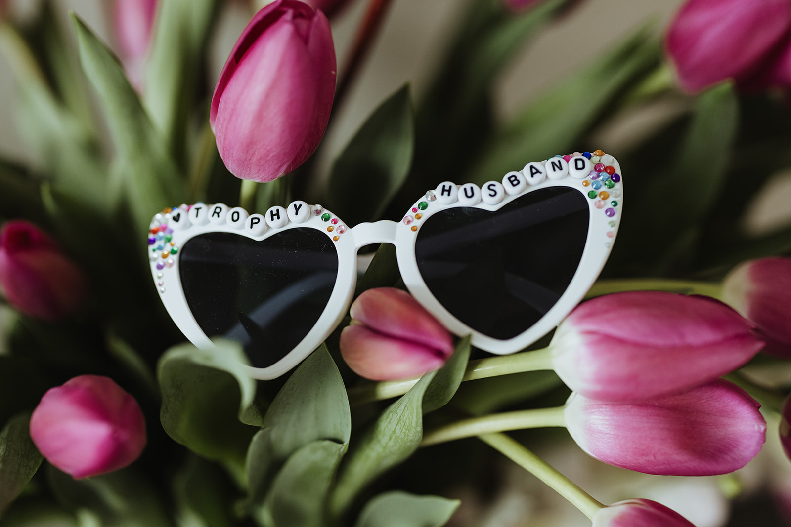 bedazzled heart shaped glasses that say "trophy husband" with pink tulips in the background