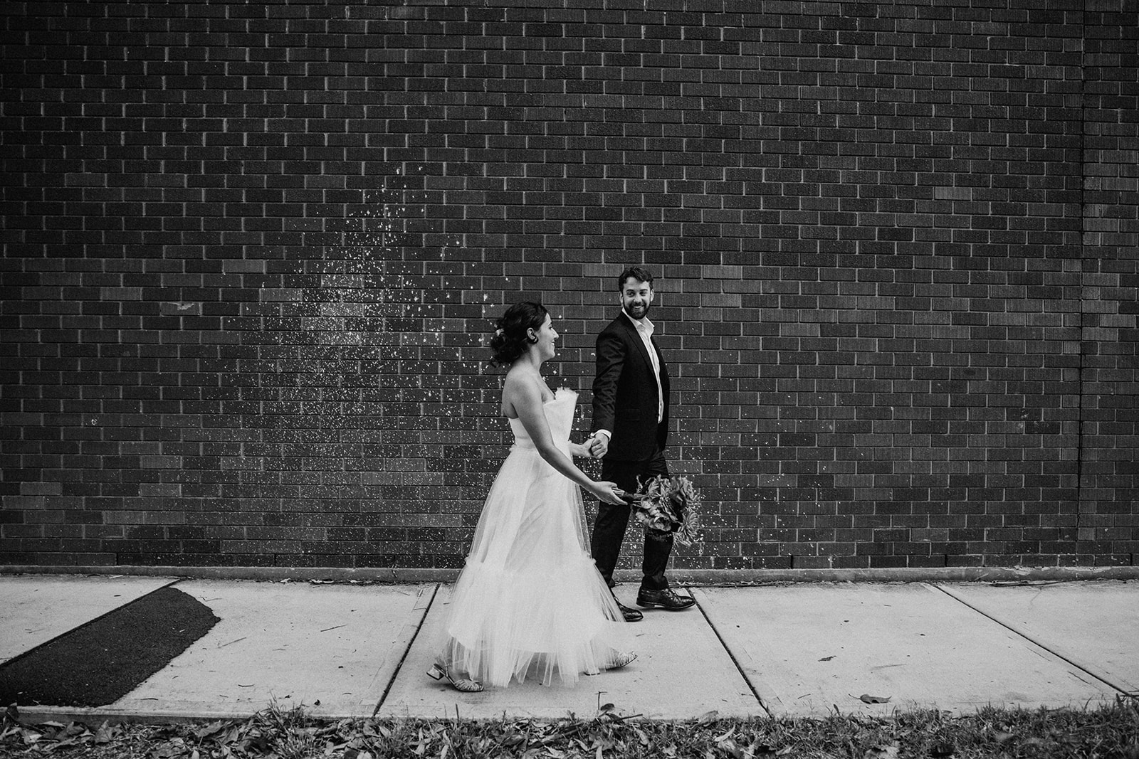 Scott Surplice bride and groom walk past a brick wall with white speckled paint